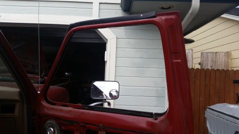 1985 Ford Bronco - The driver's side door of a red Ford Bronco is open, revealing the interior and side mirror. The vehicle is parked outside a garage with a…