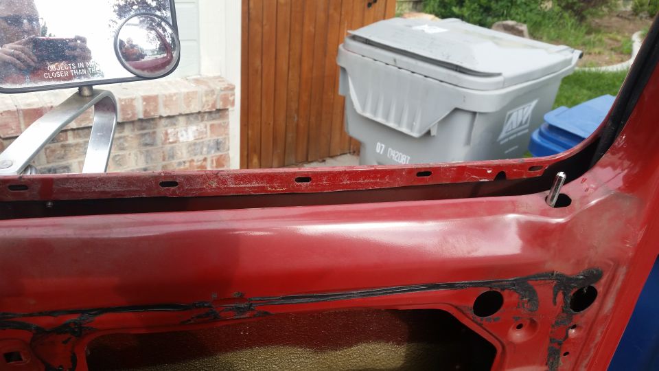 Red interior door panel with mounting holes and a metal pin, with trash bins and a wooden door in the background.