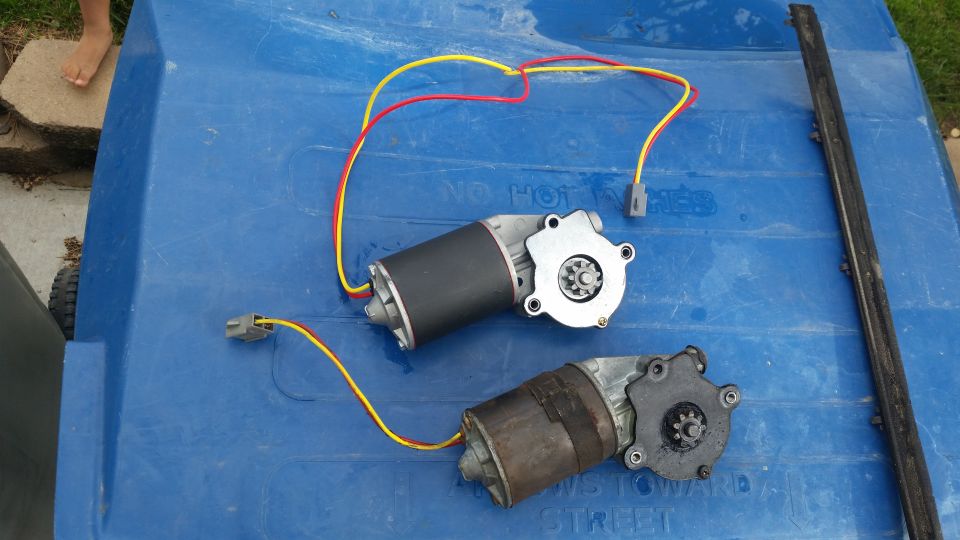 Two power window motors with wires on a blue surface next to a black trim piece.
