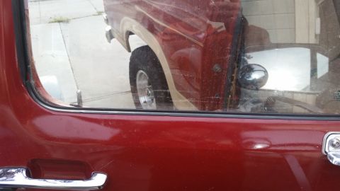 1985 Ford Bronco - Close-up view of the driver's side door and window of a red vehicle, reflecting the exterior. The reflection shows a tan stripe along the s…