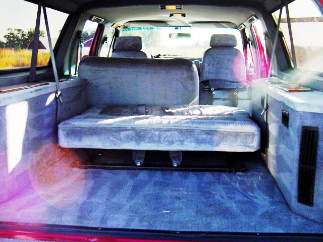 Gray bench and captain's seats over blue carpet in SUV interior, sunlit rear cargo view