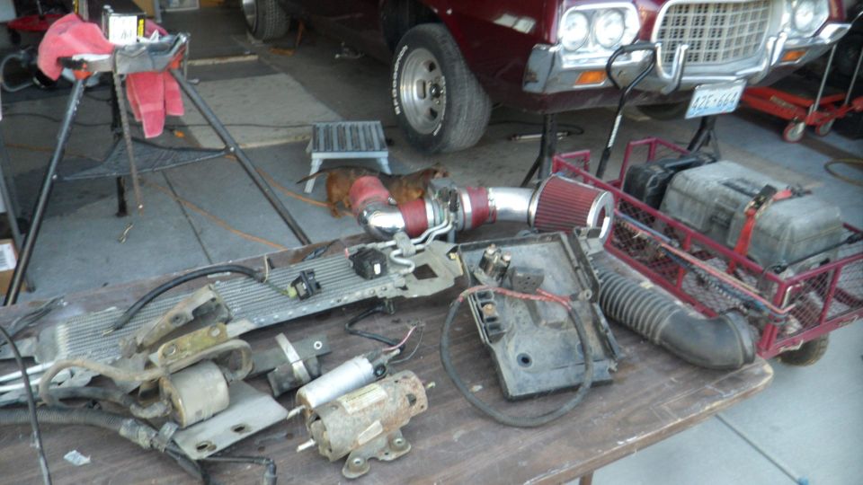 Car parts including turbo, radiator, and battery box on a table in front of a classic car.