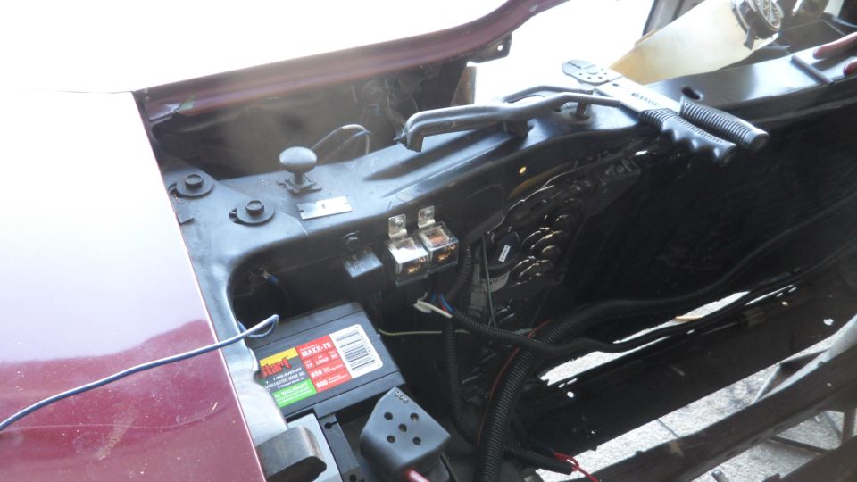 Under the hood of a vehicle showing battery, relays, wiring, and tools.