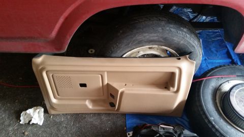 1985 Ford Bronco - A tan interior door panel is laid out on a blue tarp next to a red vehicle's fender and a tire. The door panel has a speaker grille and a h…