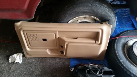 1985 Ford Bronco - A tan interior door panel is laid out on the ground next to a large tire and part of a red vehicle body. The door panel has a speaker grill…