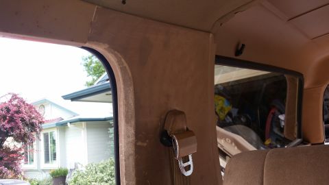 1985 Ford Bronco - Interior view of a vehicle showing a tan headliner and pillar trim, a seatbelt mechanism, and a window looking out onto a house and trees.