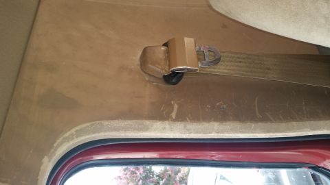 1985 Ford Bronco - A close-up view of a tan seatbelt and buckle mechanism mounted on the interior paneling of a vehicle, above a red window frame.
