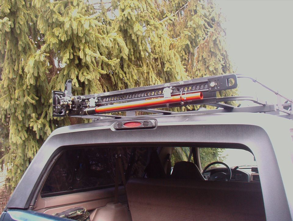Hi-Lift jack mounted on an SUV roof rack beside evergreen trees
