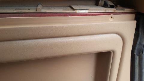 1985 Ford Bronco - A close-up view of the interior door panel of a vehicle, showing tan textured plastic with a recessed area and a visible screw hole near th…