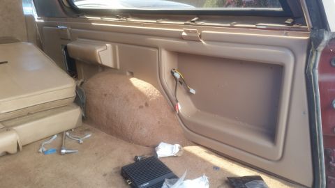 1985 Ford Bronco - Interior view of a tan Ford Bronco showing the rear passenger side panel, seat, and carpet. Wires and an amplifier are visible on the floor.