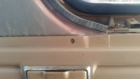 1985 Ford Bronco - A close-up view of the interior door panel of a vehicle, showing a small drilled hole and a metal door handle.