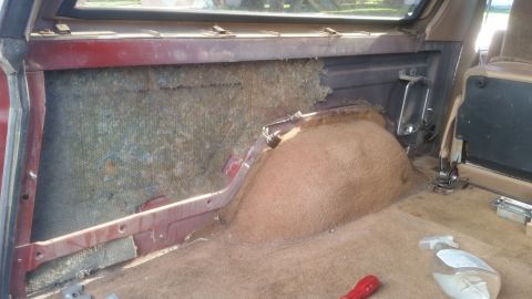 1985 Ford Bronco - The interior of a vehicle shows the rear passenger side, with exposed insulation on the left and the wheel well covered in tan carpet on th…