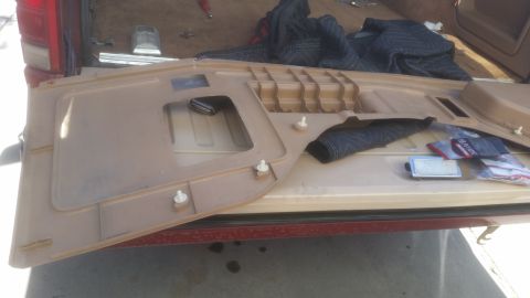 1985 Ford Bronco - A tan interior door panel is laid out on the tailgate of a vehicle, with a black blanket and some plastic-wrapped items nearby. The panel h…