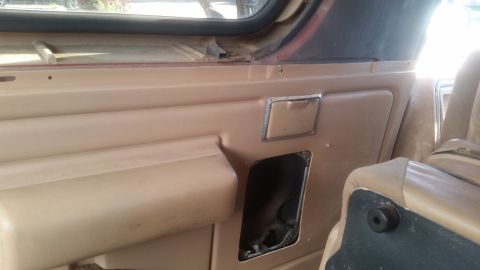 1985 Ford Bronco - Interior view of a tan door panel with a missing section revealing wiring and a chrome pull handle. A portion of a tan seat is visible on t…