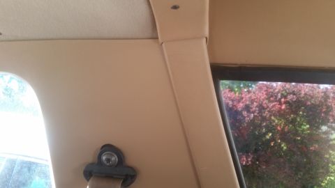 1985 Ford Bronco - Interior view of a tan-colored vehicle, showing a seatbelt buckle mechanism and a portion of the window with foliage visible outside.