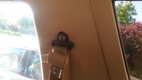 1985 Ford Bronco - Interior view of a tan seatbelt mechanism attached to the pillar of a vehicle. The seatbelt is a light tan color and is secured by a black…