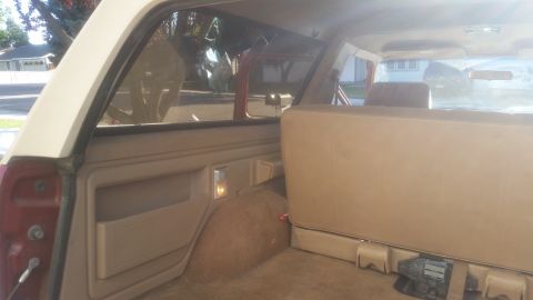 1985 Ford Bronco - The interior of a vintage Ford Bronco, showing the rear bench seat and side paneling. The exterior is visible through the rear windows, wit…