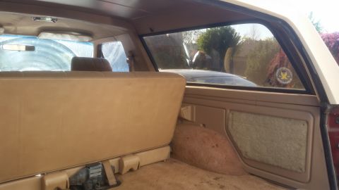 1985 Ford Bronco - Interior view of the rear of a tan SUV, showing the back seat and cargo area. A sticker is visible on the rear passenger window.