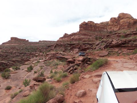 1984 Ford Bronco - Two SUVs navigate a challenging off-road trail through a desert landscape with prominent red rock formations.