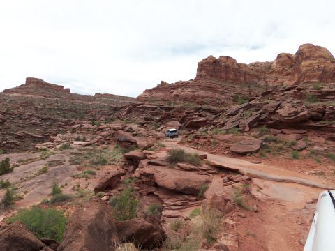 1984 Ford Bronco - An off-road vehicle navigates a path amidst towering red rock formations under a cloudy sky.