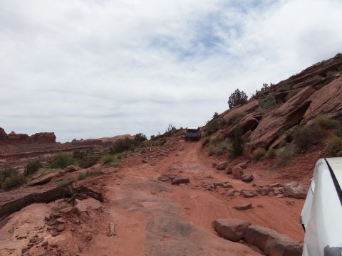 1984 Ford Bronco - A white vehicle's front corner is visible on the right, with a dark SUV navigating a rocky, uphill dirt path under a cloudy sky.