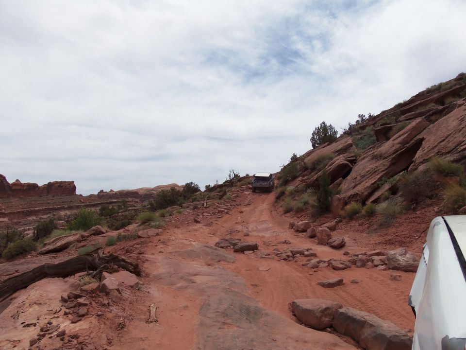 White vehicle's front corner on right, dirt road ahead with a dark SUV driving uphill