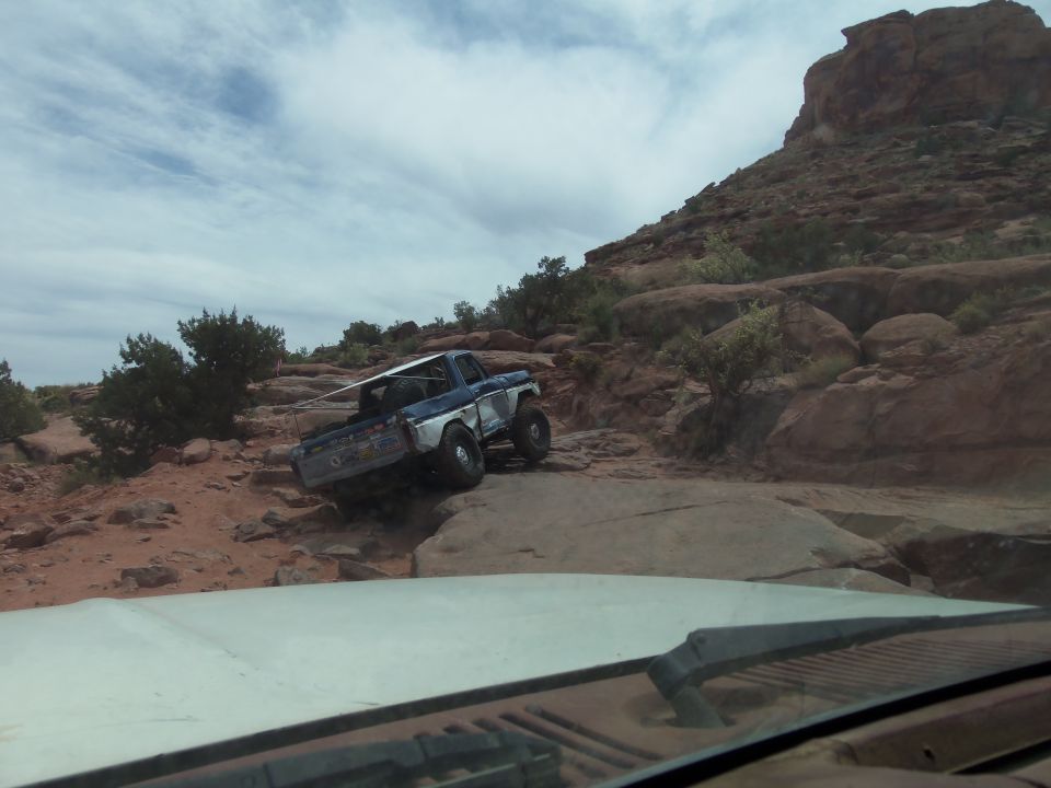 Blue and white Ford pickup truck navigating a rocky off-road trail with a large rock formation in the background