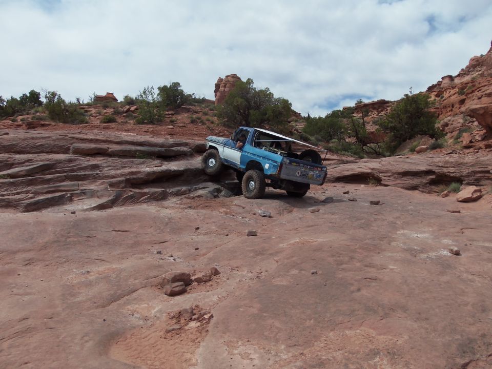 Blue and white SUV climbing a rocky incline. It has a spare tire mounted on the back and a white canopy over the bed