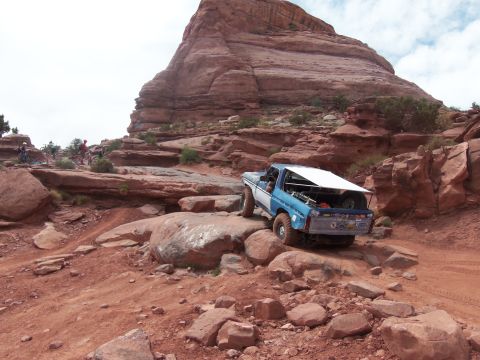 1984 Ford Bronco - This SUV navigates a rocky trail in a desert landscape, with a prominent red rock formation overhead.