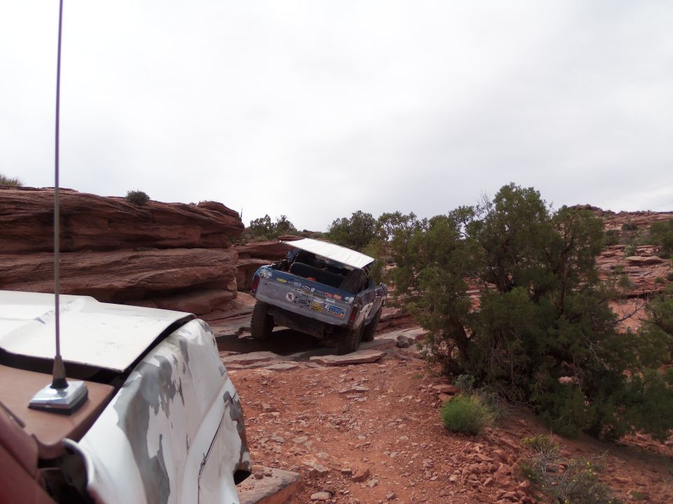 Blue off-road SUV with white roof panel, rear view, on rocky trail. Another vehicle in foreground
