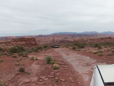 1984 Ford Bronco - Off-road vehicles navigate a desert trail with red rock formations and distant mountains under a cloudy sky.