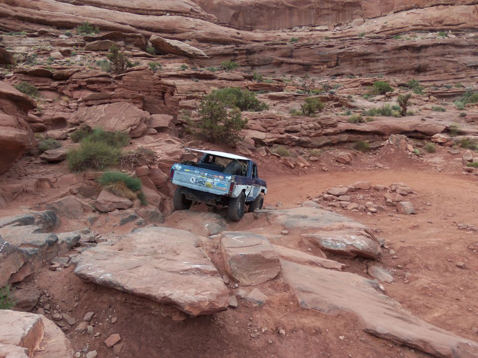 Modified 1978 Ford Bronco climbing over large rocks on a dirt trail with red rock formations in the background