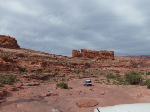 1984 Ford Bronco - Off-road vehicles navigate a dusty trail amidst the dramatic red rock formations of a desert canyon.