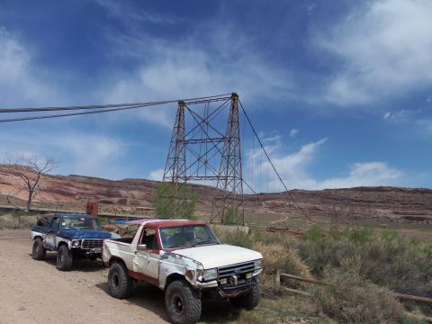 1984 Ford Bronco - Two Ford Broncos are parked on a dirt road with a large metal tower and bridge in the background.