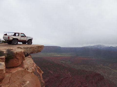 1984 Ford Bronco - A Ford Bronco sits precariously on the edge of a cliff with a vast canyon landscape in the background.