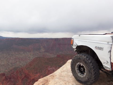 1984 Ford Bronco - A white Ford F-250 truck with large off-road tires is parked on the edge of a cliff, overlooking a vast canyon.