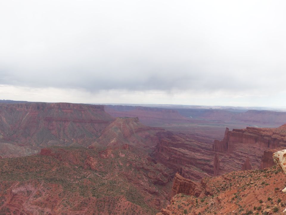Canyon landscape with red rock formations under a cloudy sky. A rock ledge is visible on the right