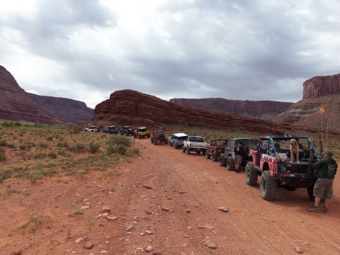 1984 Ford Bronco - A convoy of off-road vehicles travels down a dusty canyon road under a cloudy sky.