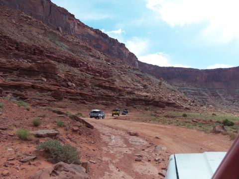 1984 Ford Bronco - Off-road vehicles navigate a dirt path in a vast canyon landscape under a partly cloudy sky.