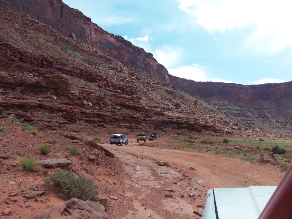 Several off-road vehicles drive on a dirt road through a canyon with red rock cliffs