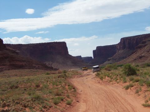 1984 Ford Bronco - Two SUVs navigate a dusty dirt road winding through a canyon under a bright blue sky.
