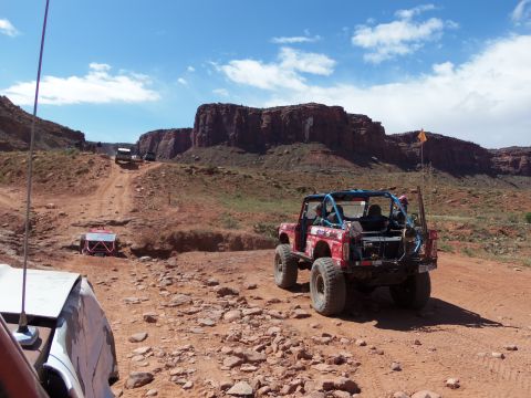 1984 Ford Bronco - Several off-road vehicles navigate a dusty canyon trail under a bright blue sky.