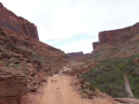 1984 Ford Bronco - A lone off-road vehicle navigates a dusty trail through a canyon landscape.