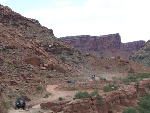 1984 Ford Bronco - Multiple off-road vehicles traverse a winding dirt path in a vast, arid canyon.