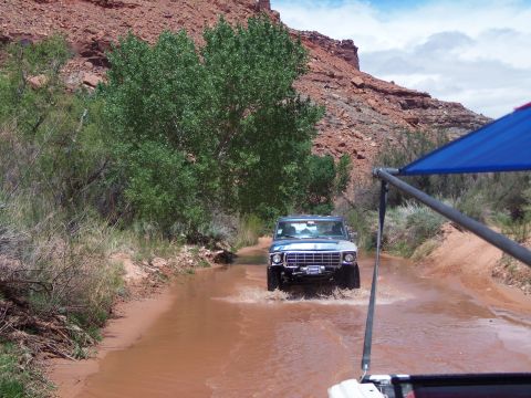 1984 Ford Bronco - A silver Ford Bronco navigates a shallow, muddy riverbed in a desert landscape with red rock formations overhead.