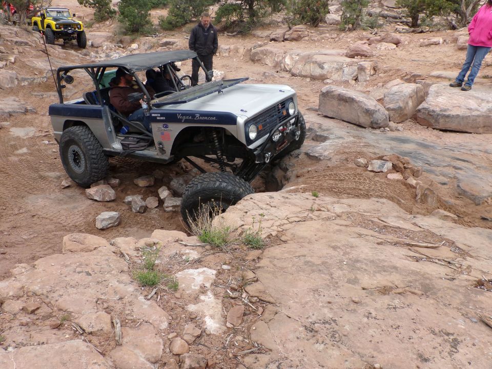 Modified off-road vehicle with roll cage navigating a rocky trail; another yellow off-road vehicle is visible in the background