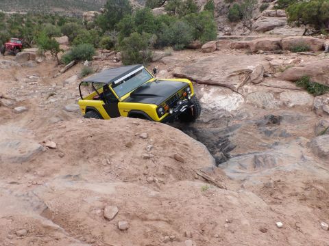 1984 Ford Bronco - A yellow and black 1984 Ford Bronco tackles a challenging off-road trail, with another red vehicle visible in the background.