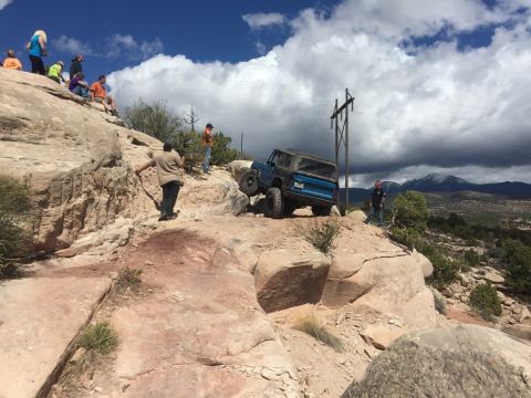1984 Ford Bronco - A blue 1984 Ford Bronco navigates a challenging rocky trail with spectators watching from a nearby ridge.