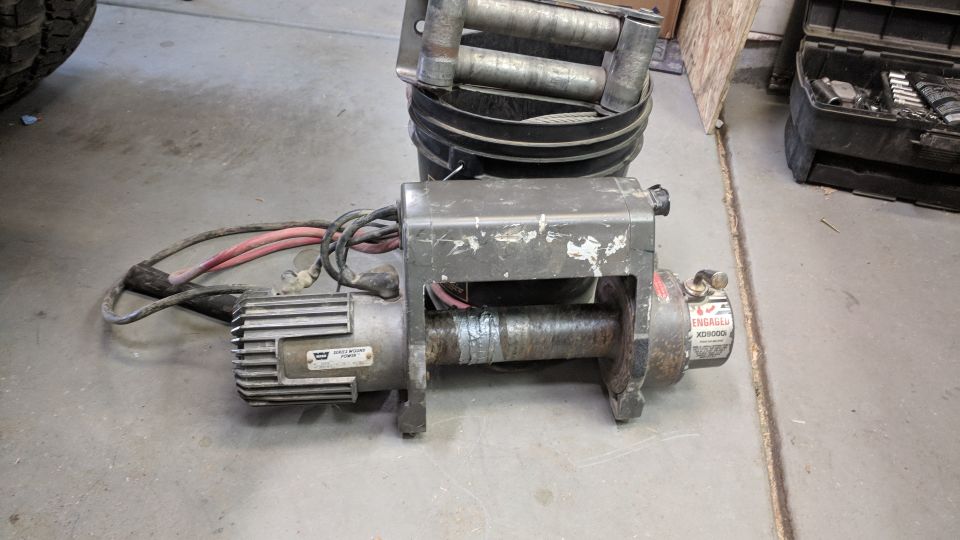 worn electric winch with red and black cables, sitting on concrete floor next to a bucket with rollers