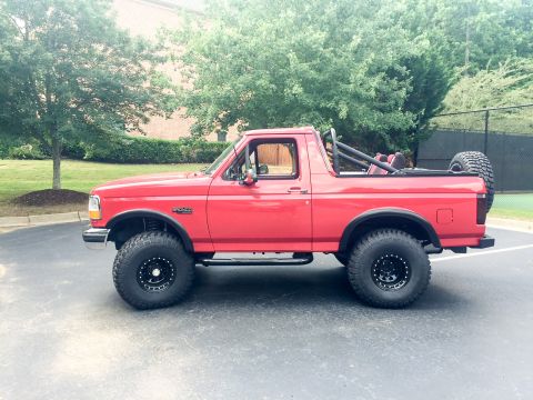 1994 Ford Bronco - A red 1994 Ford Bronco is parked outdoors, showing its side profile with off-road tires and a spare tire mounted at the rear.